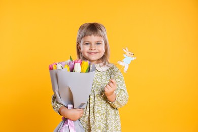 Photo of Smiling little girl with bouquet of tulips and decorative Easter bunny on orange background