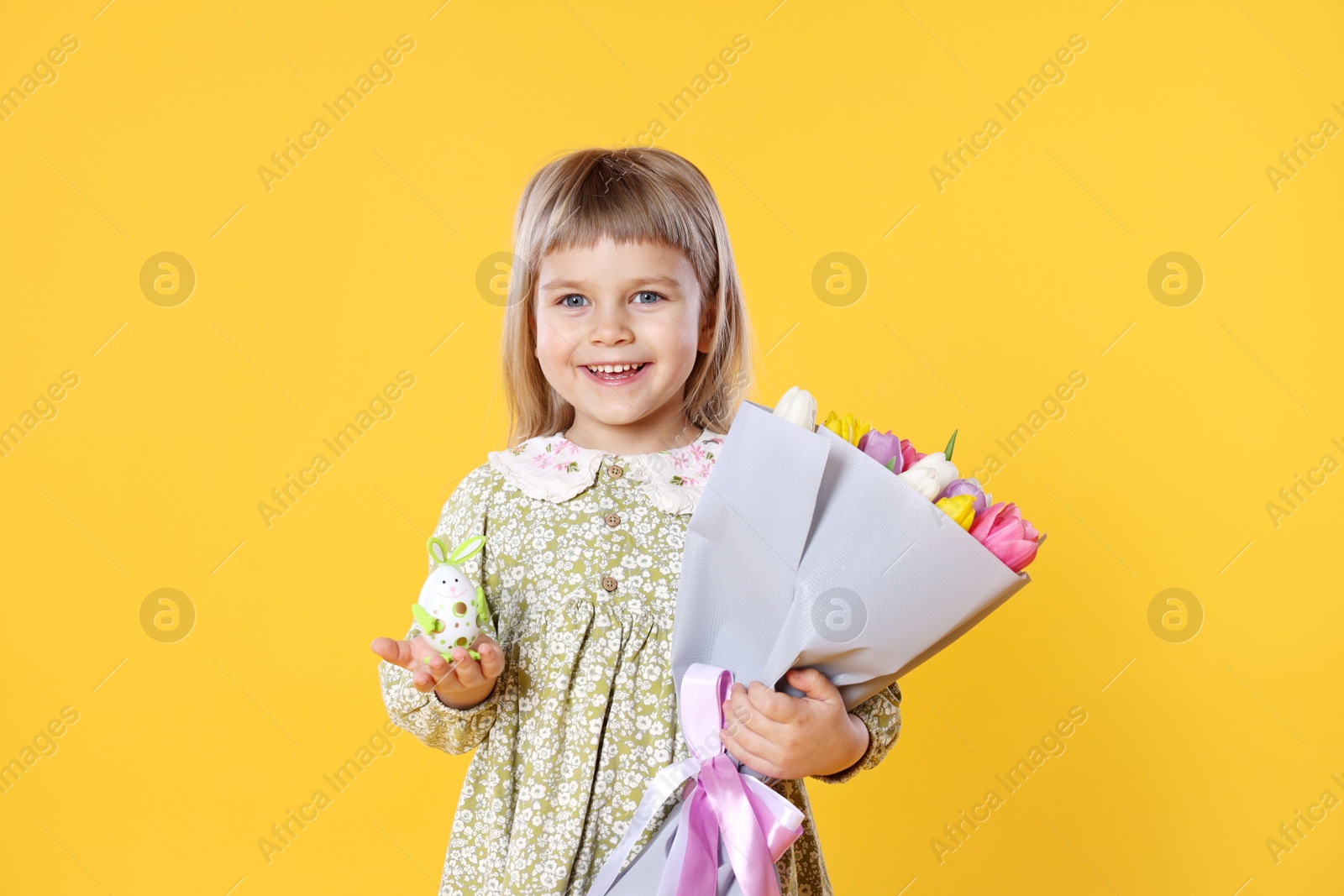 Photo of Smiling little girl with bouquet of tulips and decorative Easter egg on orange background