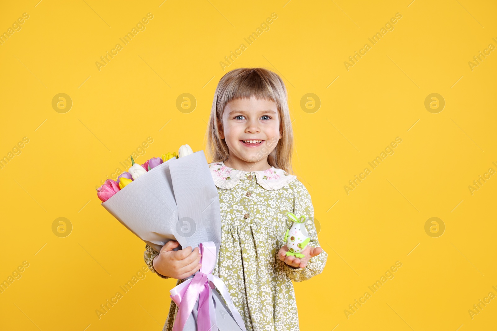 Photo of Smiling little girl with bouquet of tulips and decorative Easter egg on orange background