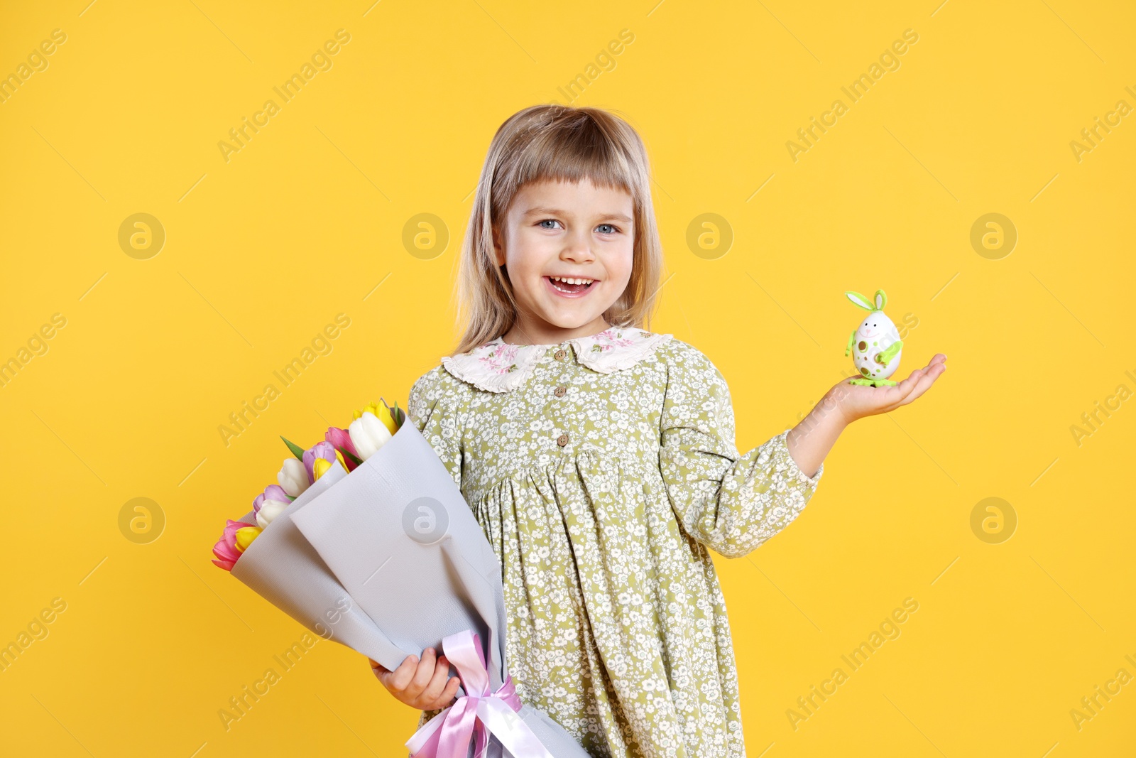 Photo of Smiling little girl with bouquet of tulips and decorative Easter egg on orange background