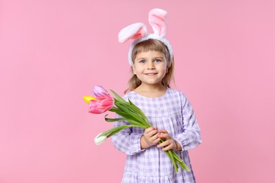 Photo of Smiling little girl in headband with bunny ears holding bouquet of tulips on pink background, space for text. Easter celebration