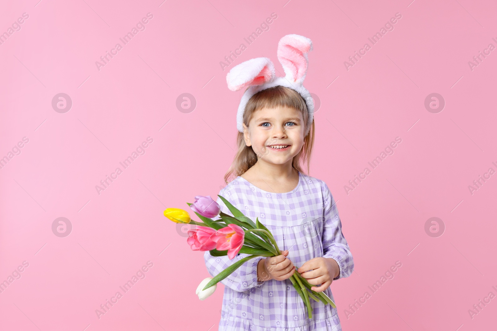 Photo of Smiling little girl in headband with bunny ears holding bouquet of tulips on pink background, space for text. Easter celebration