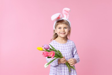 Smiling little girl in headband with bunny ears holding bouquet of tulips on pink background, space for text. Easter celebration