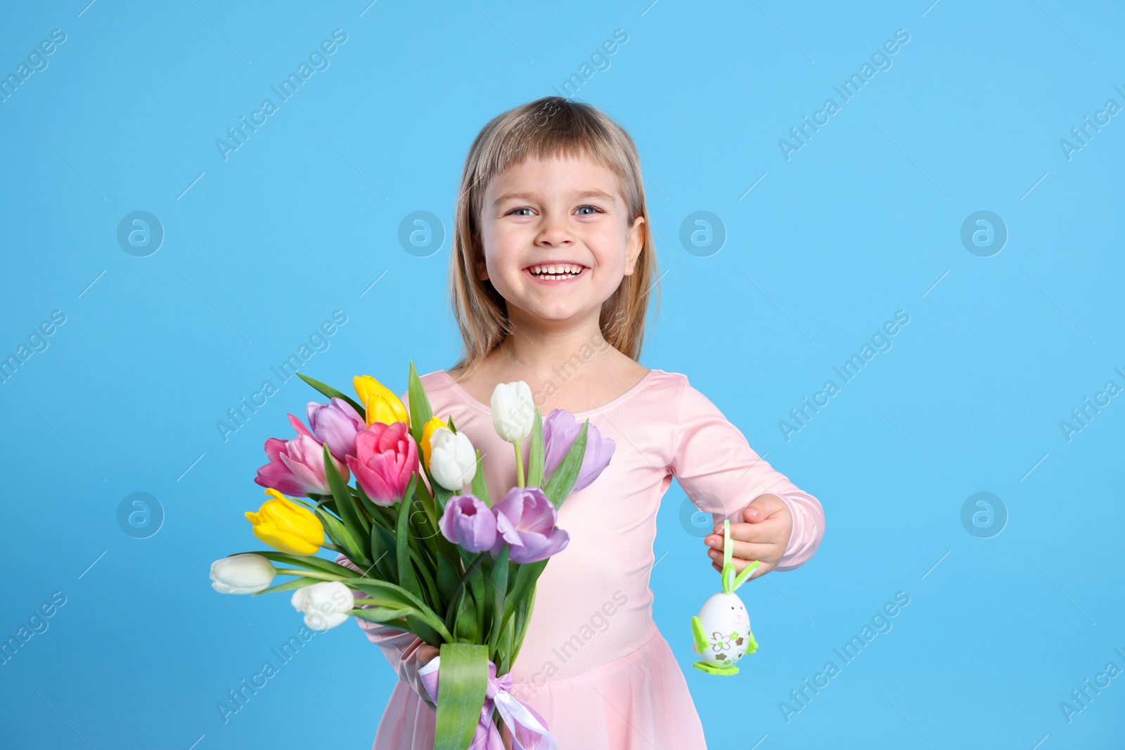 Photo of Smiling little girl with bouquet of tulips and decorative Easter egg on light blue background