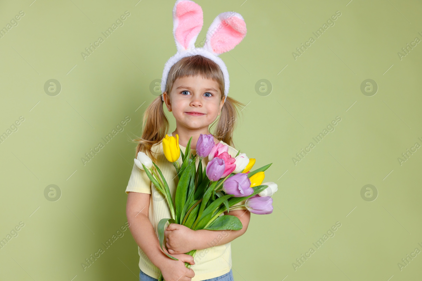 Photo of Cute little girl in headband with bunny ears holding bouquet of tulips on green background. Easter celebration