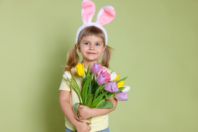 Photo of Cute little girl in headband with bunny ears holding bouquet of tulips on green background. Easter celebration