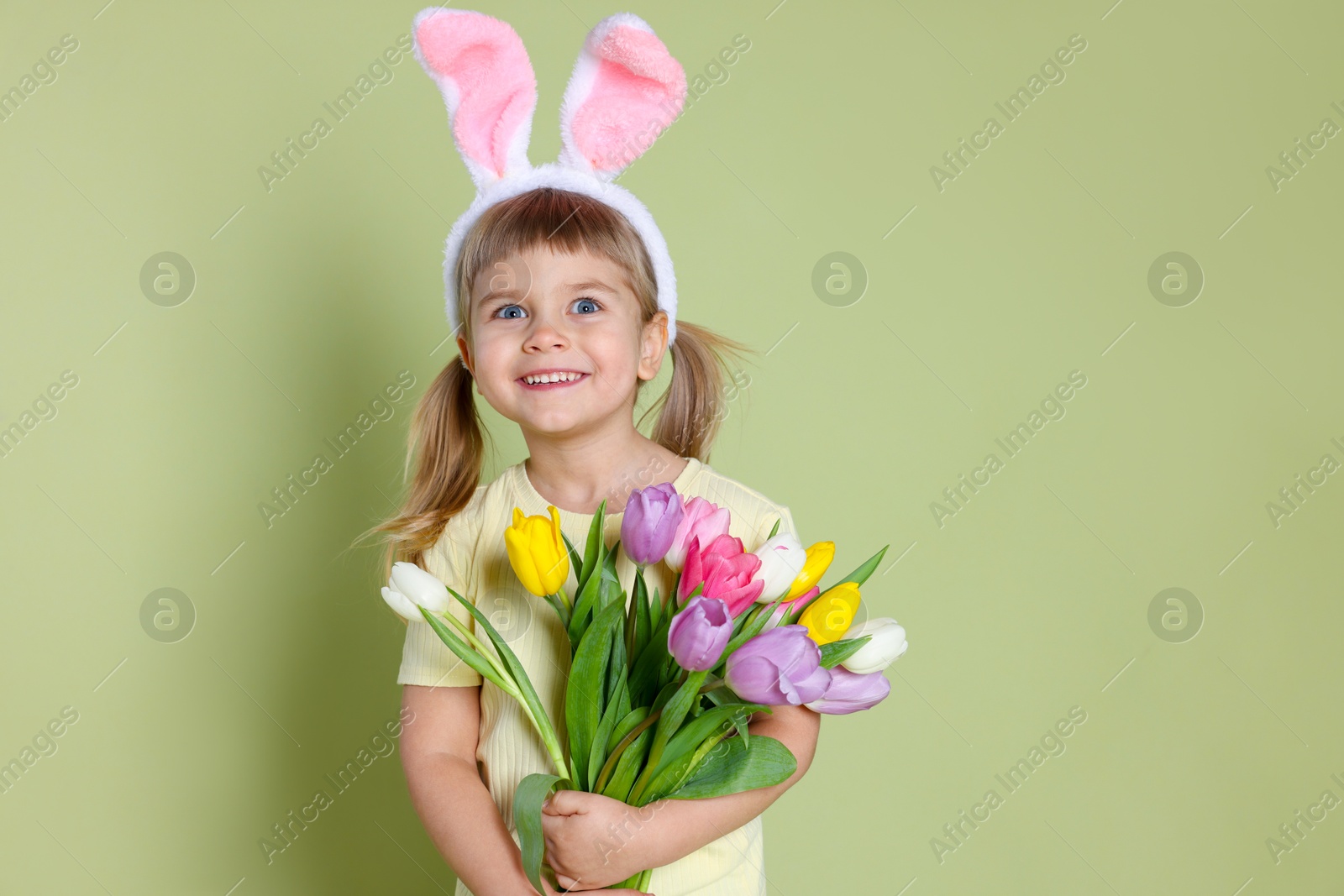 Photo of Happy little girl in headband with bunny ears holding bouquet of tulips on green background. Easter celebration