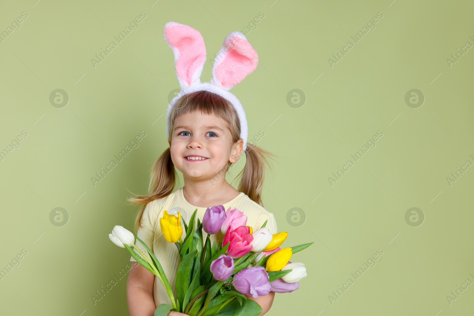 Photo of Happy little girl in headband with bunny ears holding bouquet of tulips on green background. Easter celebration