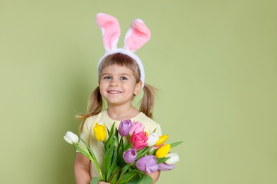 Happy little girl in headband with bunny ears holding bouquet of tulips on green background. Easter celebration