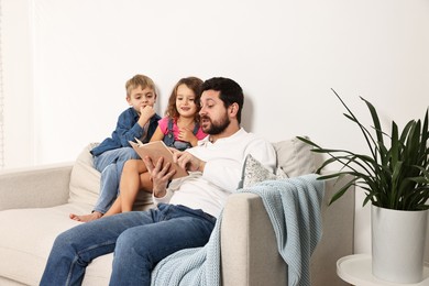 Photo of Overwhelmed father reading book to his children on sofa at home