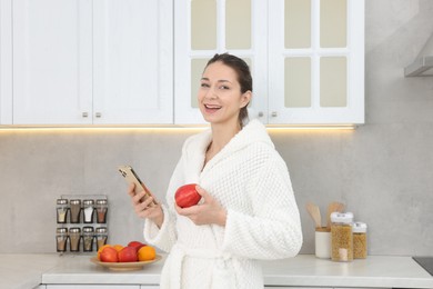 Photo of Beautiful woman with apple using smartphone after spa procedure in kitchen