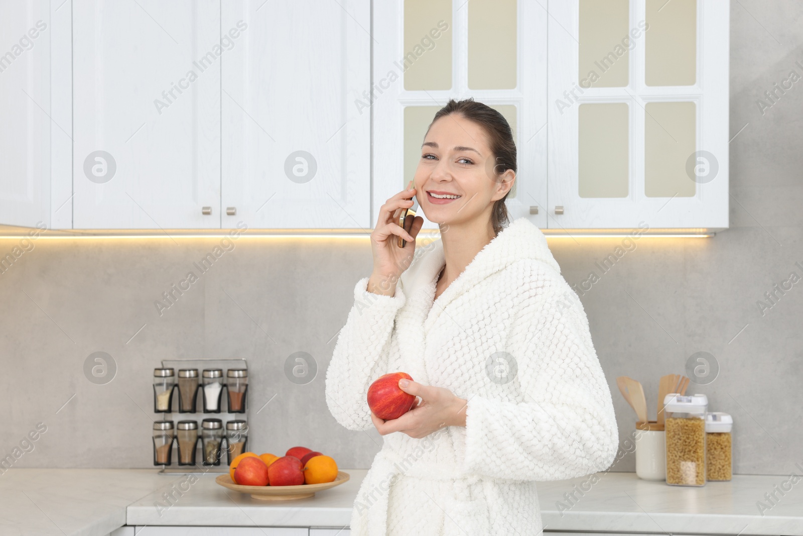 Photo of Beautiful woman with apple talking on smartphone after spa procedure in kitchen