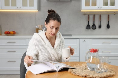 Photo of Beautiful woman with cup of drink reading magazine after spa procedure at wooden table in kitchen