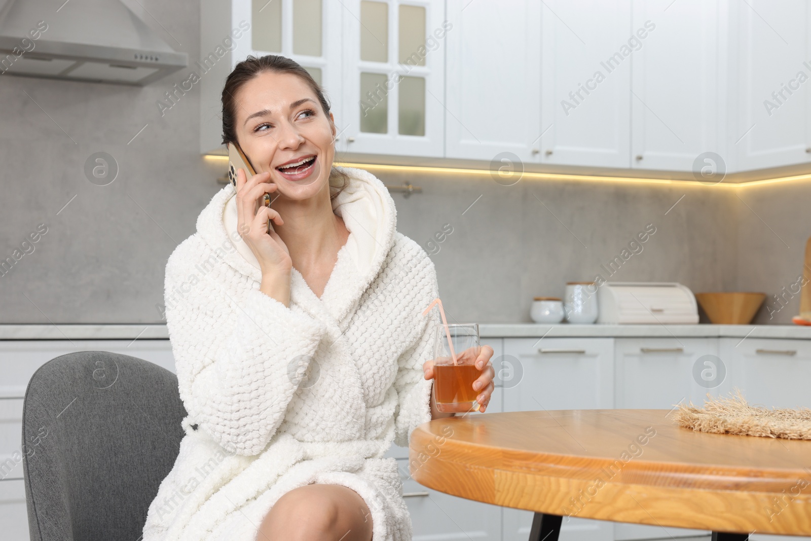 Photo of Beautiful woman with glass of juice talking on smartphone after spa procedure in kitchen