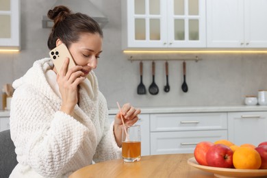 Photo of Beautiful woman with glass of juice talking on smartphone after spa procedure in kitchen