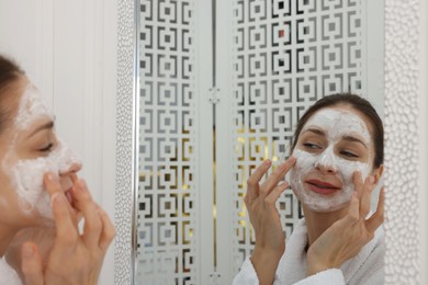 Photo of Spa day. Beautiful woman applying mask onto her face near mirror indoors