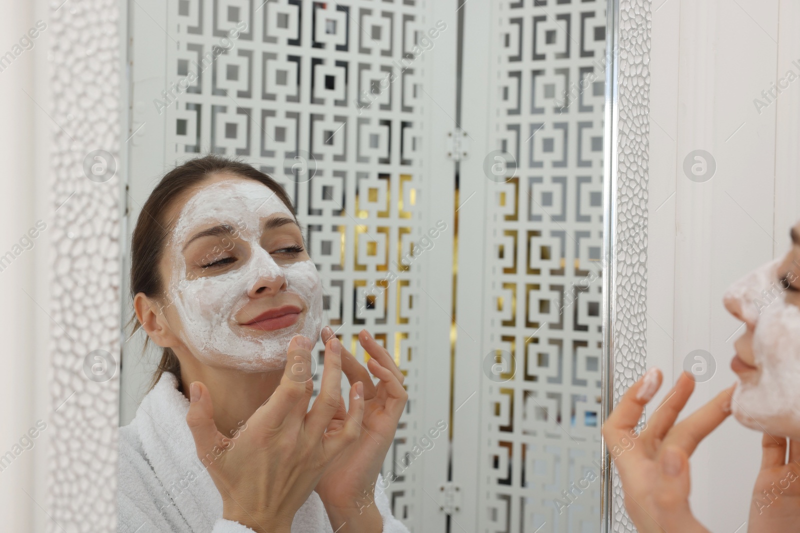 Photo of Spa day. Beautiful woman applying mask onto her face near mirror indoors