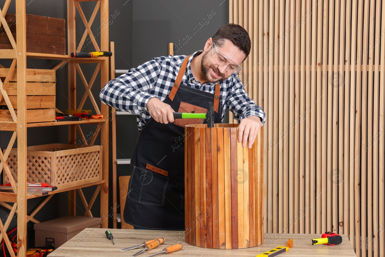 Photo of Relaxing hobby. Smiling man repairing wooden trash can with hammer at table in workshop