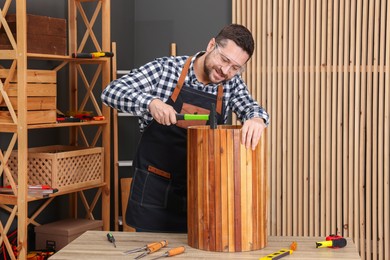 Photo of Relaxing hobby. Smiling man repairing wooden trash can with hammer at table in workshop