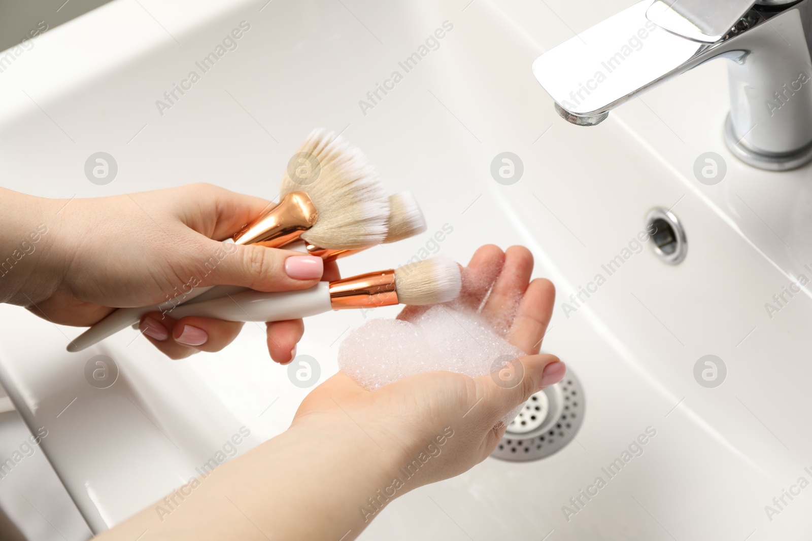 Photo of Woman washing makeup brushes with soap over sink, closeup