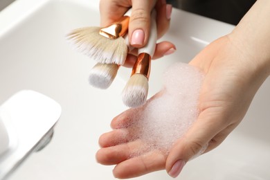 Photo of Woman washing makeup brushes with soap over sink, closeup