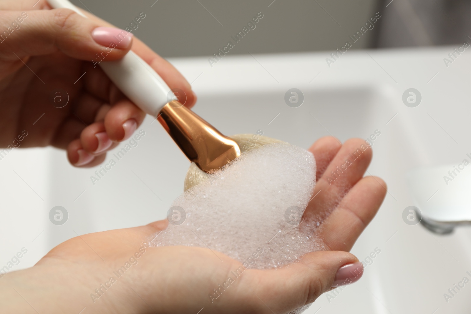 Photo of Woman washing makeup brush with soap over sink, closeup