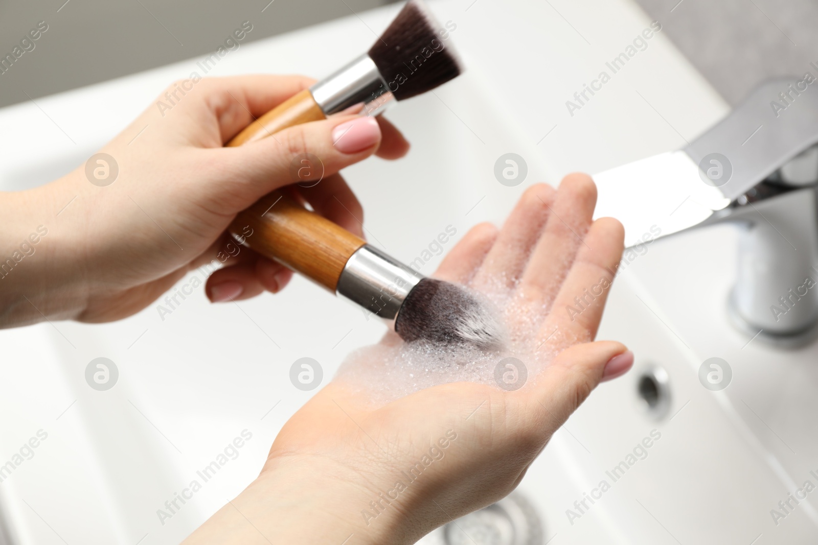 Photo of Woman washing makeup brushes with soap over sink, closeup