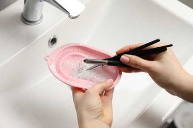 Photo of Woman washing makeup brushes with soap and cleansing pad over sink, above view