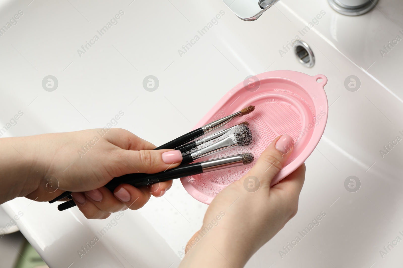 Photo of Woman washing makeup brushes with soap and cleansing pad over sink, above view