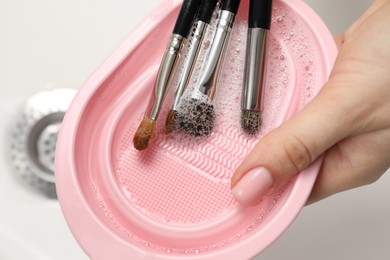 Photo of Woman washing makeup brushes with soap and cleansing pad in sink, closeup