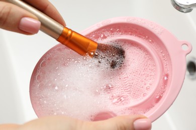 Photo of Woman washing makeup brush with soap and cleansing pad over sink, closeup