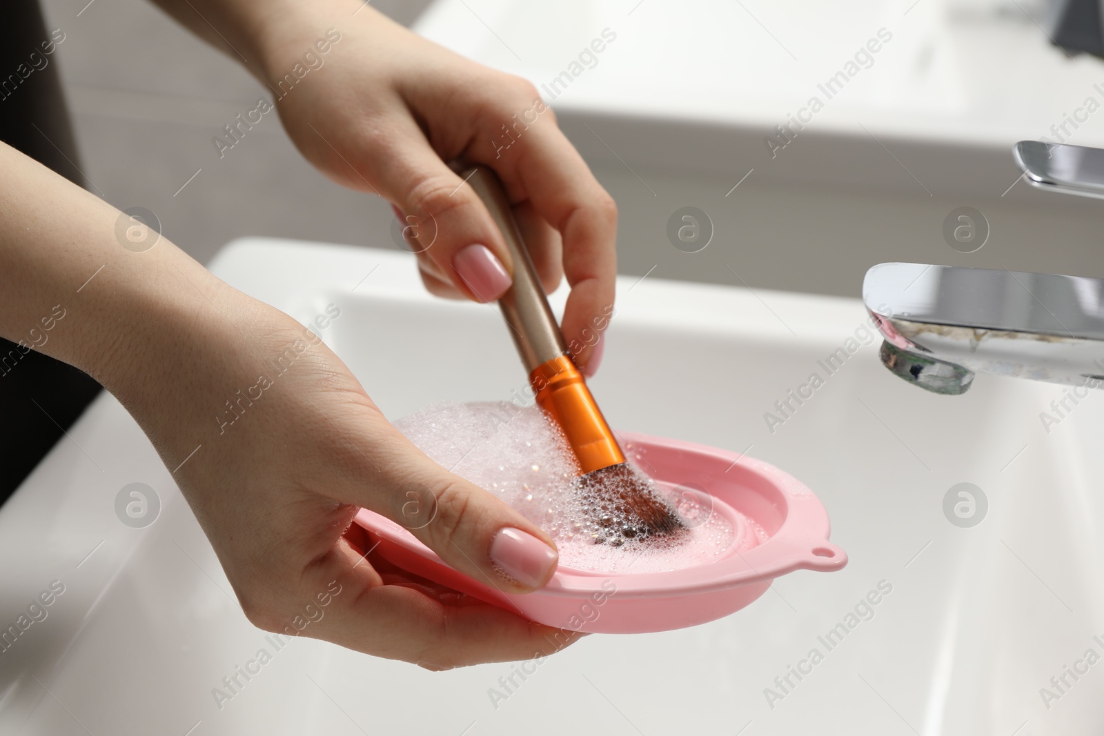 Photo of Woman washing makeup brush with soap and cleansing pad over sink, closeup