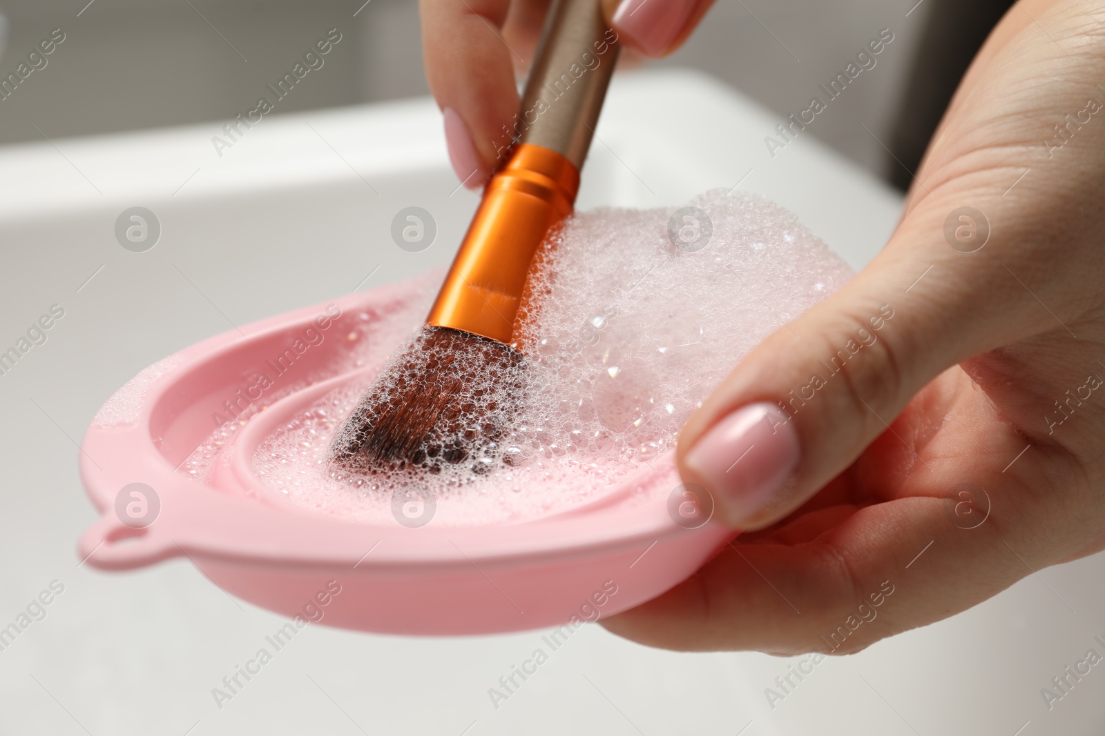 Photo of Woman washing makeup brush with soap and cleansing pad over sink, closeup