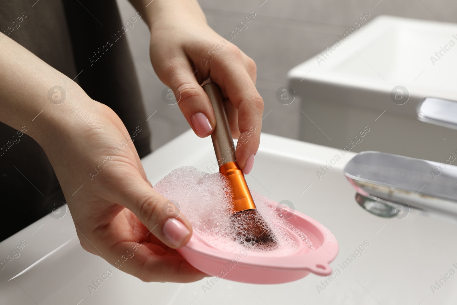 Photo of Woman washing makeup brush with soap and cleansing pad over sink, closeup