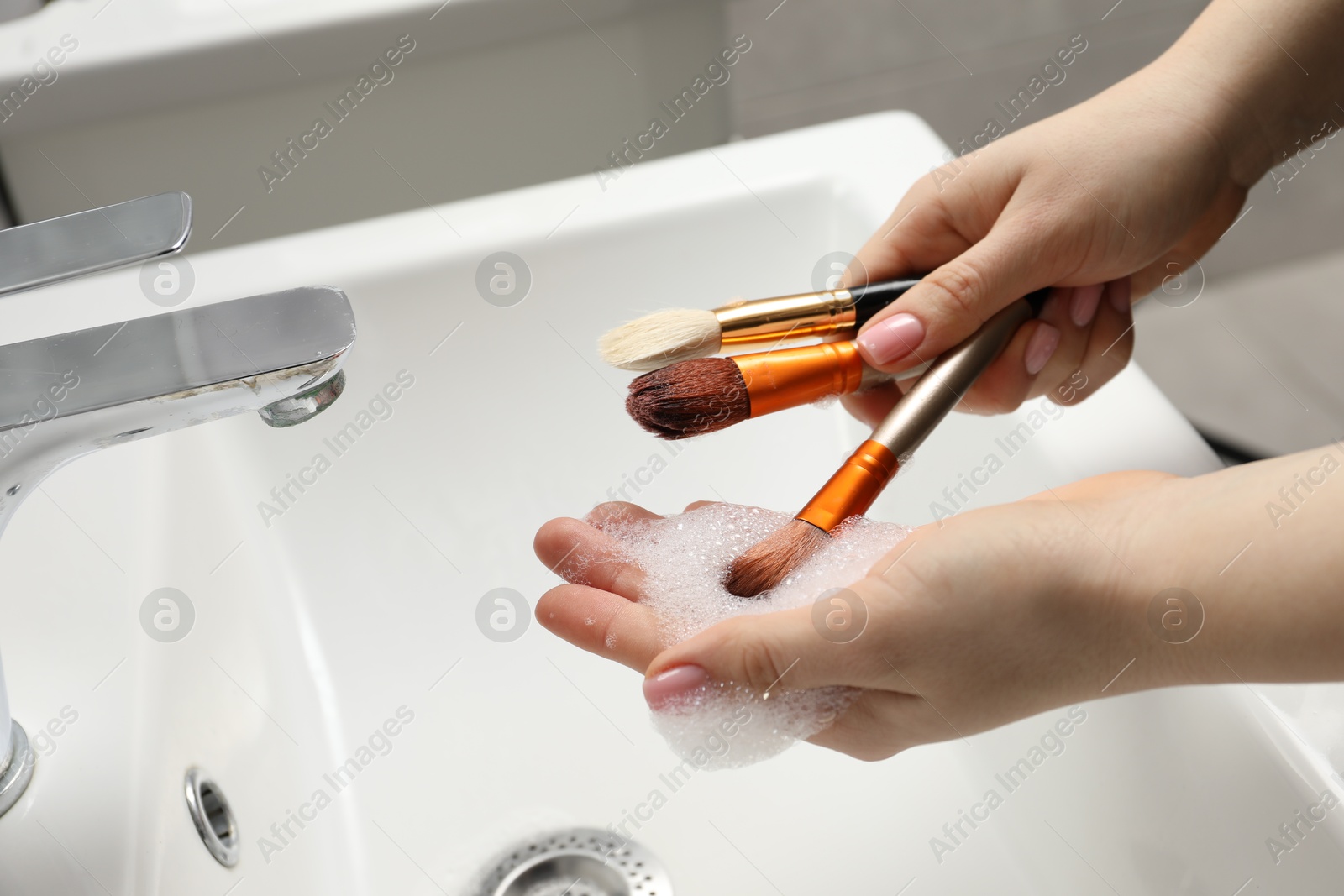 Photo of Woman washing makeup brushes with soap over sink, closeup