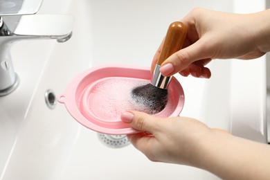 Photo of Woman washing makeup brush with soap and cleansing pad over sink, closeup