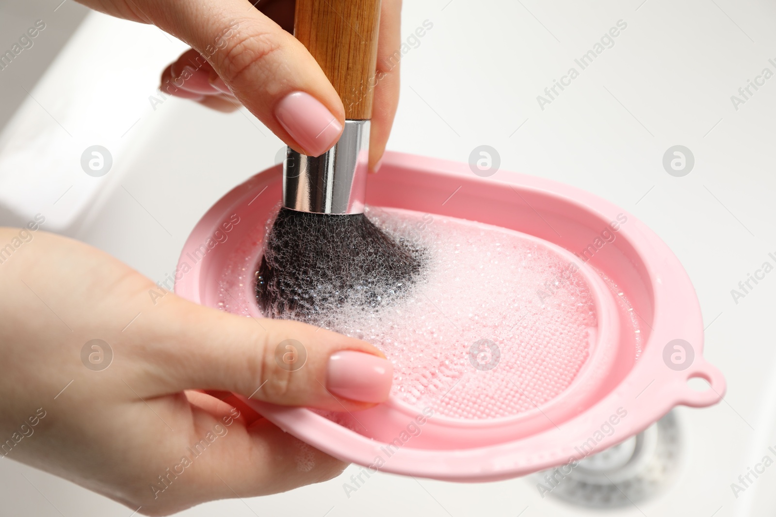 Photo of Woman washing makeup brush with soap and cleansing pad over sink, closeup