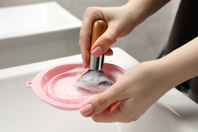 Photo of Woman washing makeup brush with soap and cleansing pad over sink, closeup