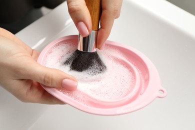 Photo of Woman washing makeup brush with soap and cleansing pad over sink, closeup