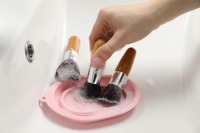 Photo of Woman washing makeup brushes with soap and cleansing pad in sink, closeup