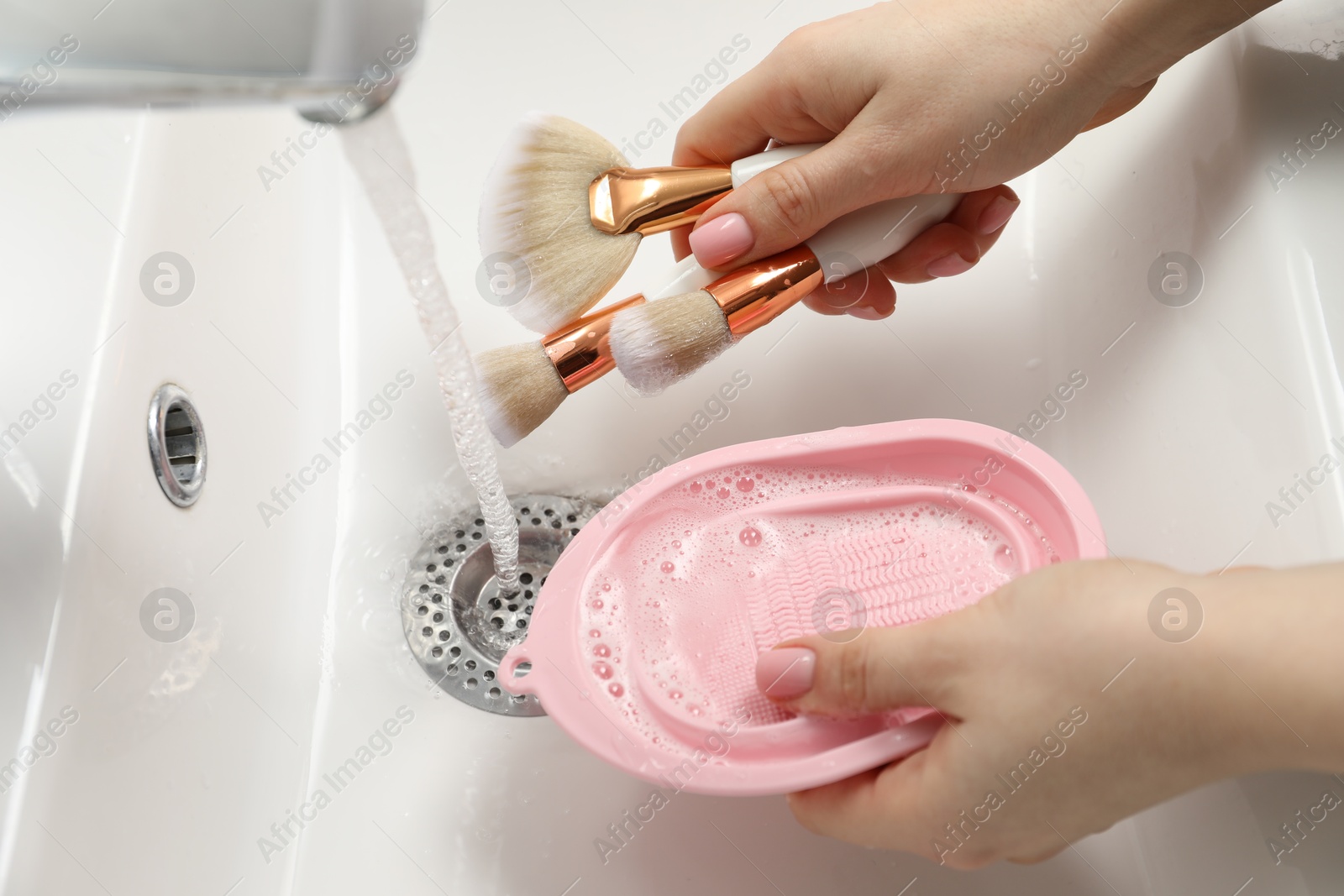 Photo of Woman with cleansing pad washing makeup brushes under stream of water in sink, closeup