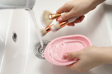 Photo of Woman with cleansing pad washing makeup brushes under stream of water in sink, closeup