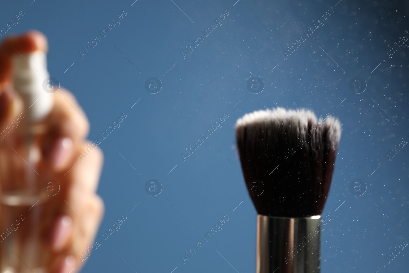 Photo of Woman cleaning makeup brush with spray on blue background, selective focus