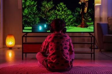 Little boy in headphones playing video game indoors at night, back view