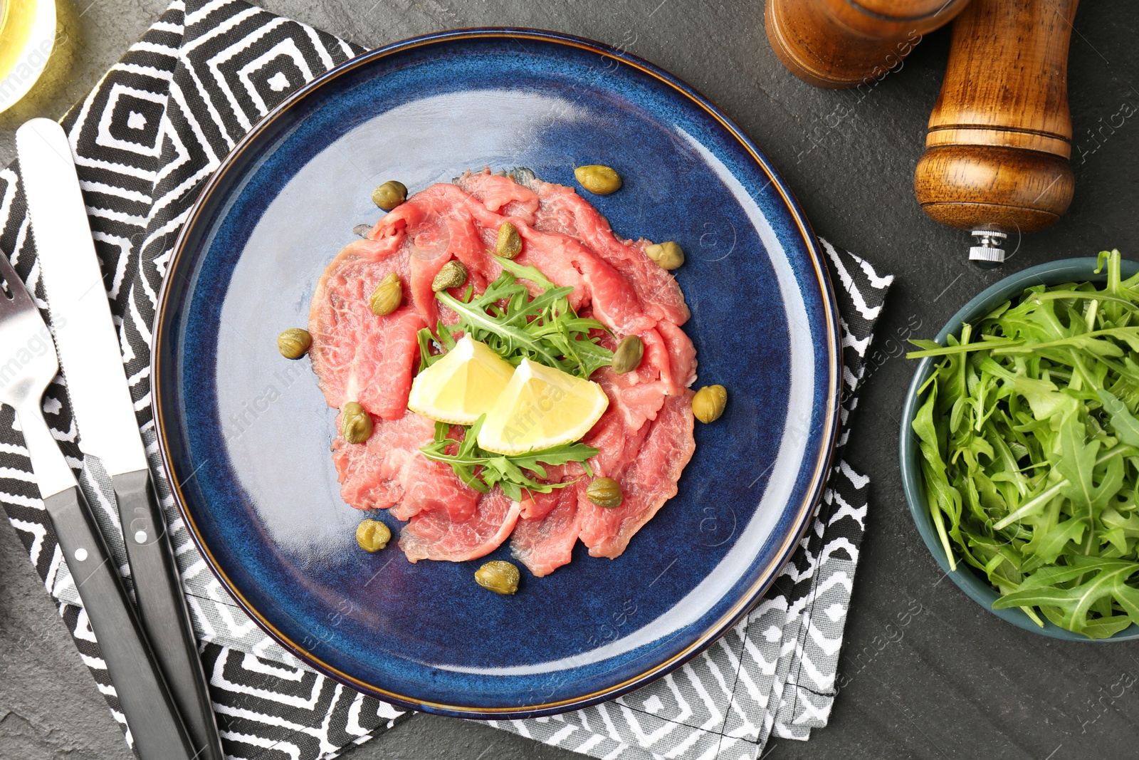 Photo of Fresh beef carpaccio with arugula, lemon and capers served on dark textured table, flat lay