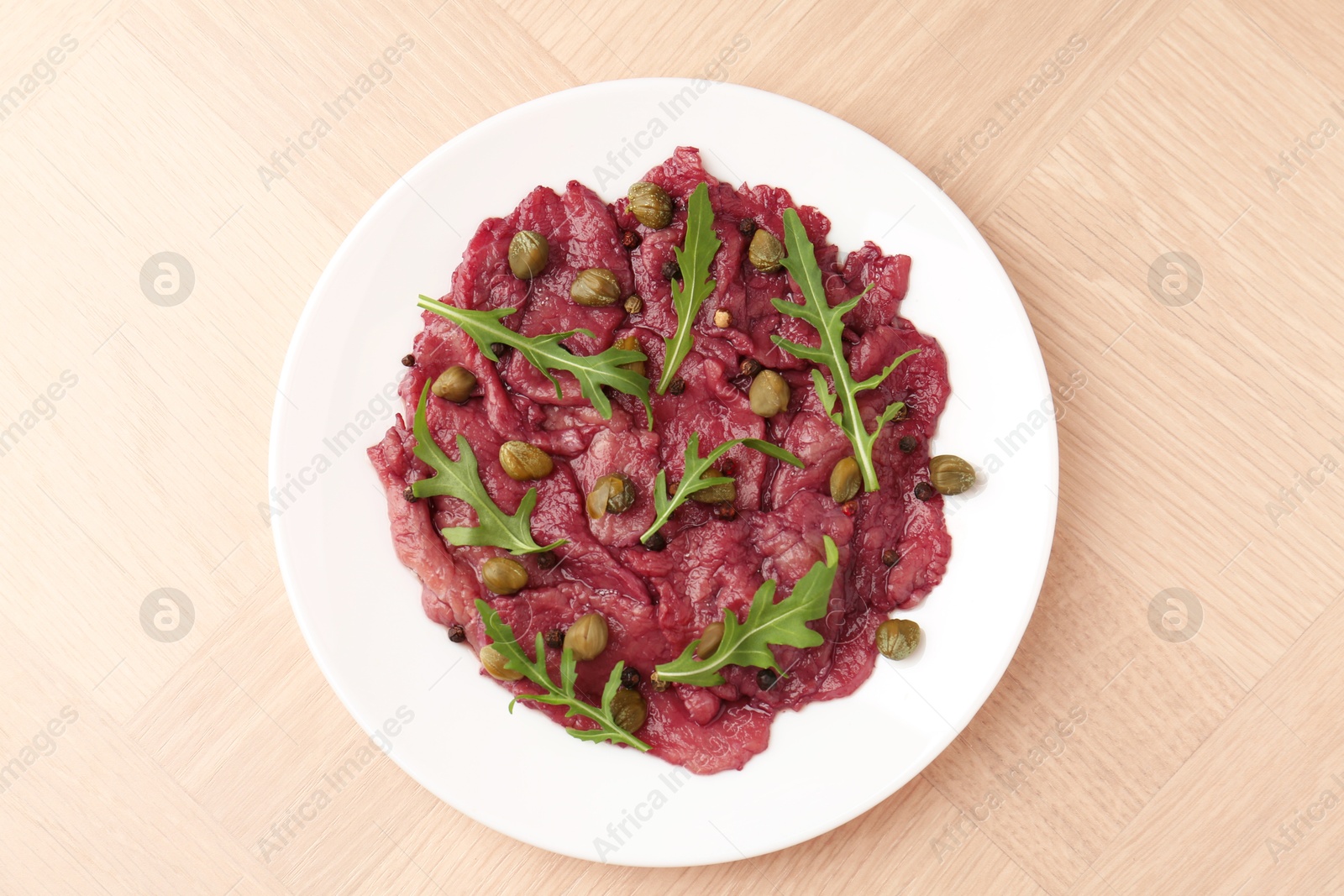 Photo of Fresh beef carpaccio with arugula and capers on wooden table, top view