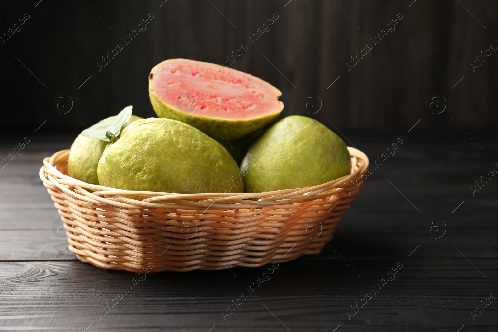 Photo of Fresh whole and cut guava fruits in wicker basket on black wooden table, closeup