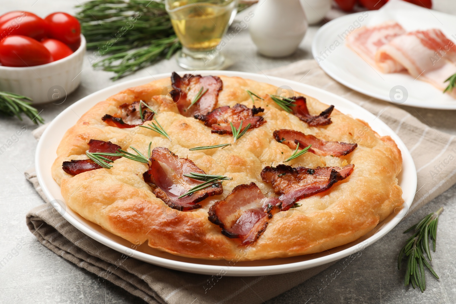 Photo of Delicious focaccia bread with bacon, rosemary, oil and tomatoes on light grey table, closeup