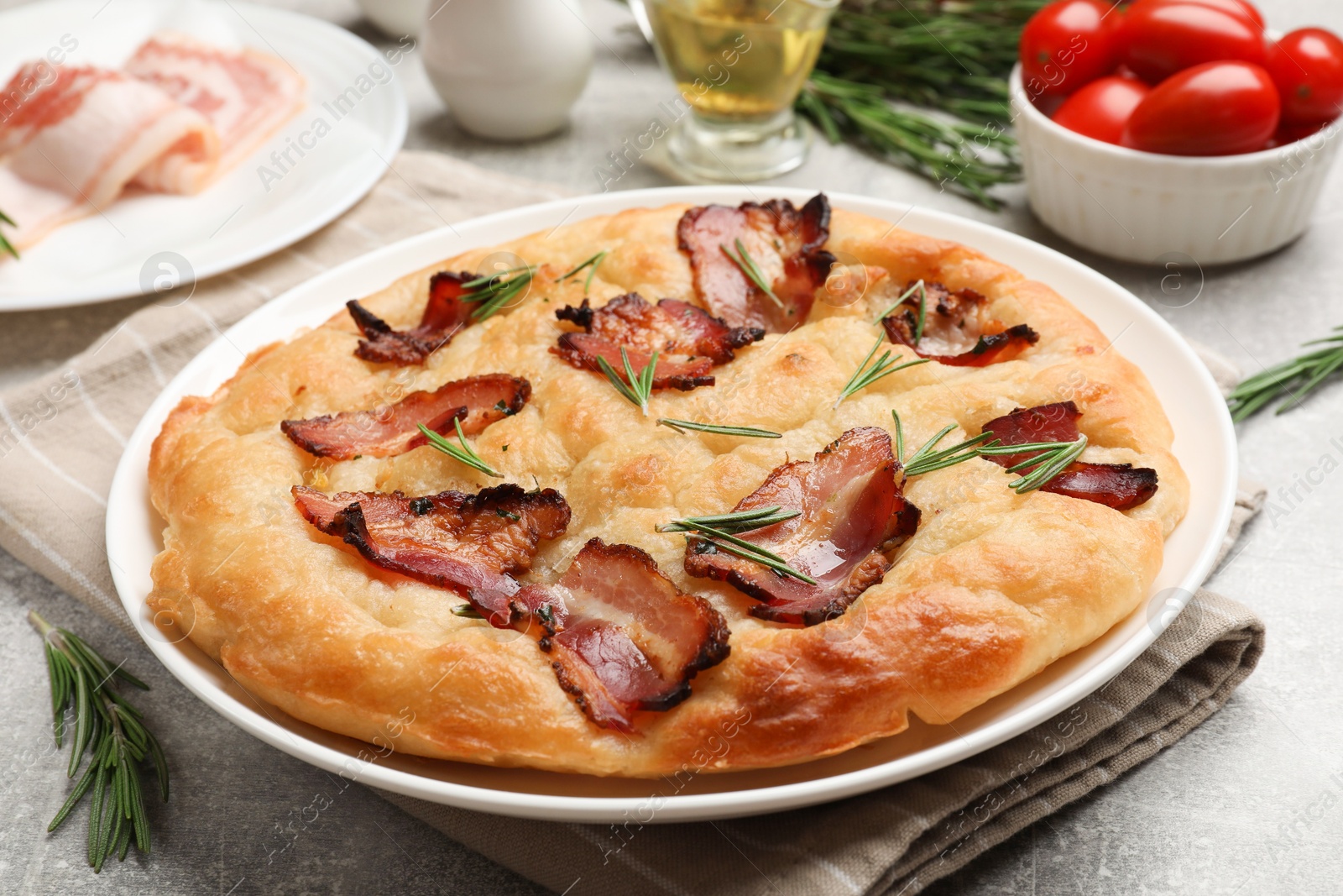 Photo of Delicious focaccia bread with bacon, rosemary, oil and tomatoes on light grey table, closeup