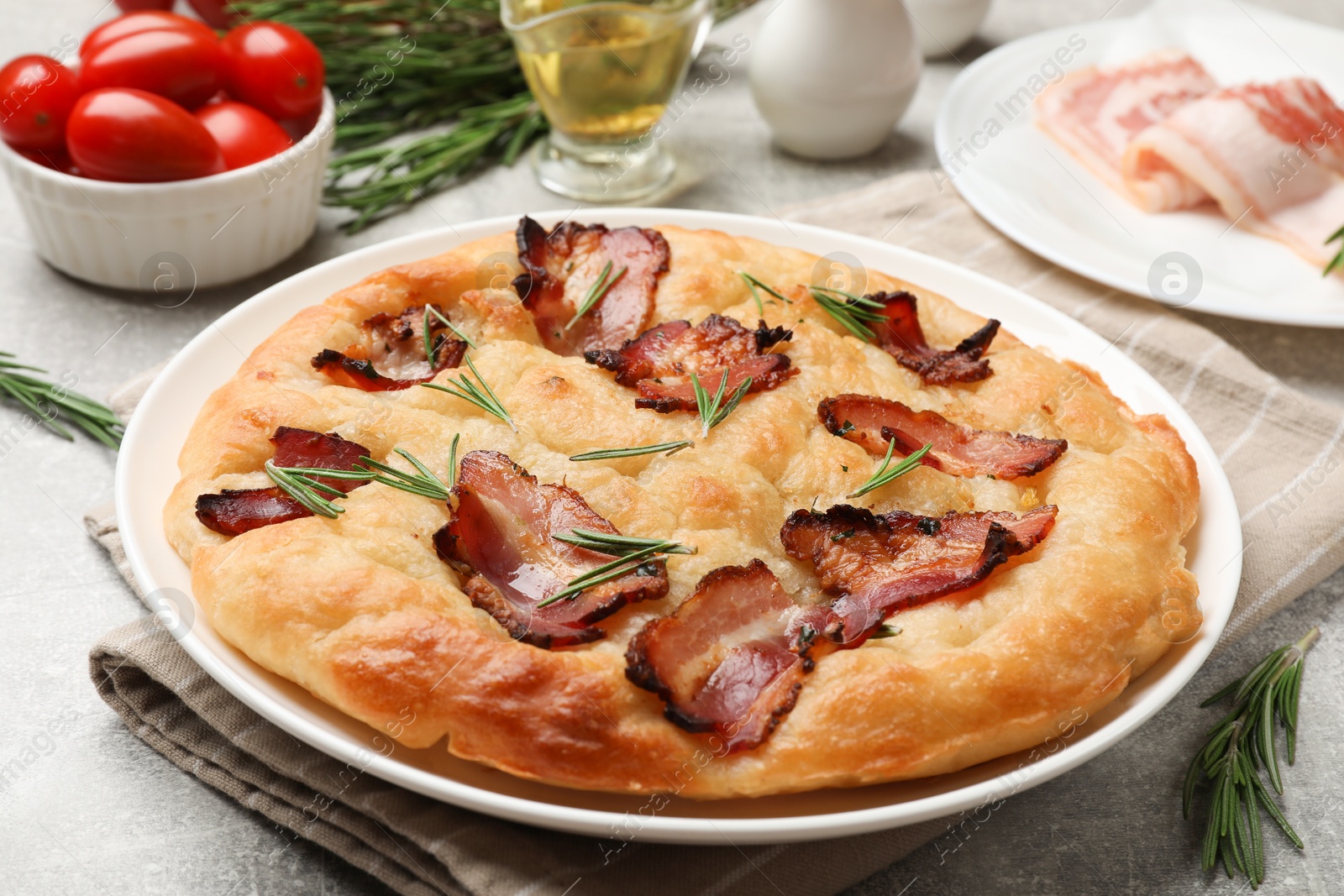 Photo of Delicious focaccia bread with bacon, rosemary, oil and tomatoes on light grey table, closeup